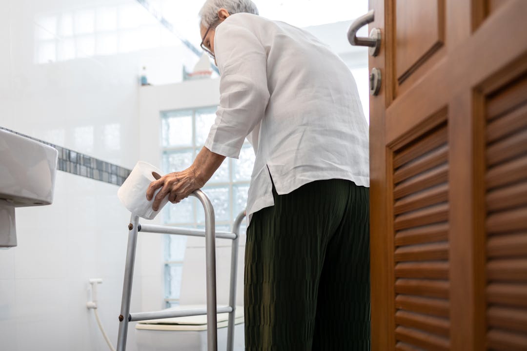 Senior person holding tissue roll near a toilet bowl, using walker to walk to the bathroom stock photo