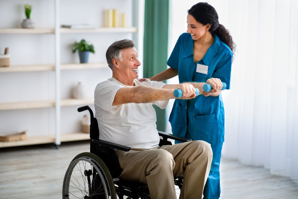 Female physiotherapist helping elderly man in wheelchair do exercises with dumbbells at health center stock photo